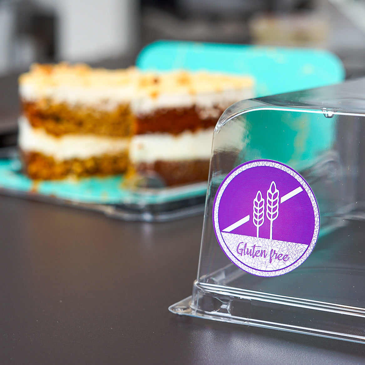 A clear plastic display cover with a vibrant purple 'Gluten Free' circular sticker attached, featuring a crossed-out wheat icon. In the blurred background, two slices of carrot cake with white frosting are visible, suggesting safe serving of allergy-friendly baked goods.