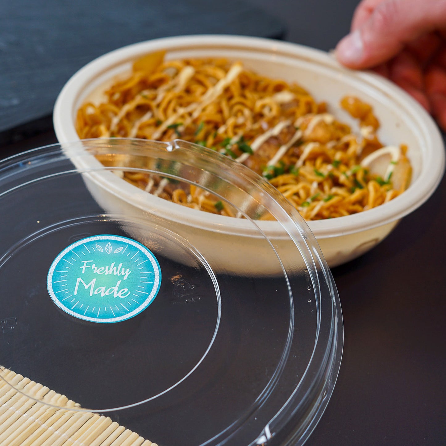 A clear plastic lid with a 'Freshly Made' circular sticker in teal, featuring a leaf design, placed in front of a bowl of noodles with sauce and garnishes. Hands are visible holding the bowl in the background, perfect for food service businesses showcasing fresh meals