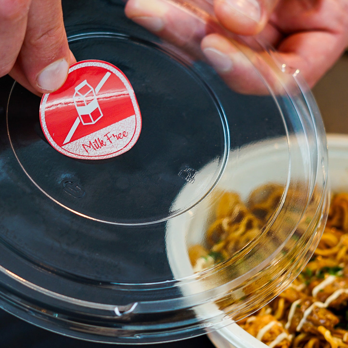 Hands placing a red circular 'Milk Free' sticker with a crossed-out milk carton icon onto a clear plastic food container lid. The lid partially covers a meal with noodles and sauce, indicating careful labeling for dietary needs.