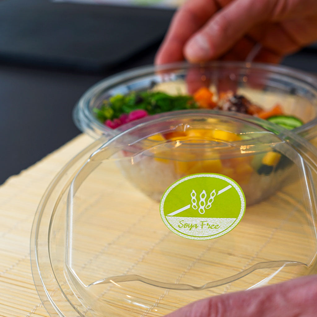 Hands placing a clear plastic lid with a green circular 'Soya Free' sticker onto a food container. The sticker features a crossed-out soybean icon, emphasizing safe packaging for those with soy allergies, with colorful food visible inside the bowl