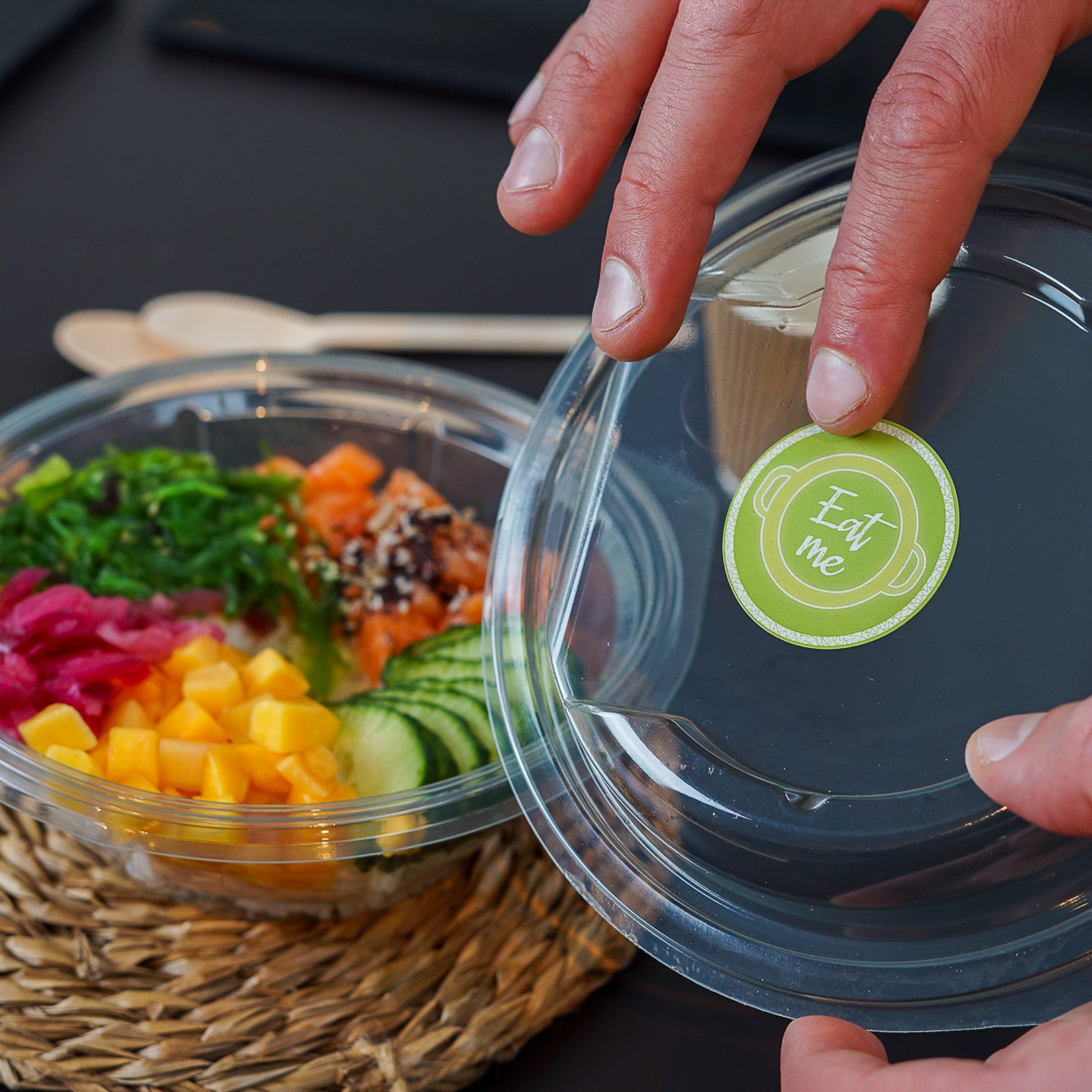 Hands holding a clear plastic lid with a green circular 'Eat me' sticker, featuring a pot icon, over a poke bowl filled with salmon, mango, cucumber, seaweed, and pickled onions. Ideal for food businesses to encourage consumption of fresh, ready-to-eat meals