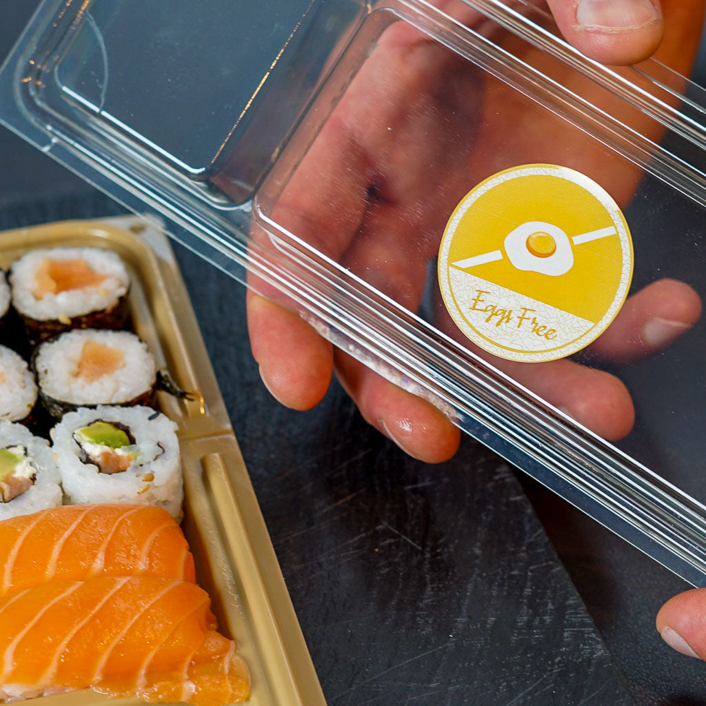 Hands holding a clear plastic food container lid with a yellow circular 'Eggs Free' sticker, featuring a crossed-out fried egg icon. A tray of sushi, including salmon nigiri and maki rolls, is visible below, highlighting safe food handling for customers with egg allergies