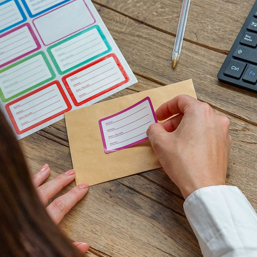 Hands peeling a purple-bordered mailing label from a sheet of colorful labels to apply it to a brown envelope. A black keyboard and pen are also visible on the rustic wooden table, ideal for showcasing easy-to-use shipping labels.