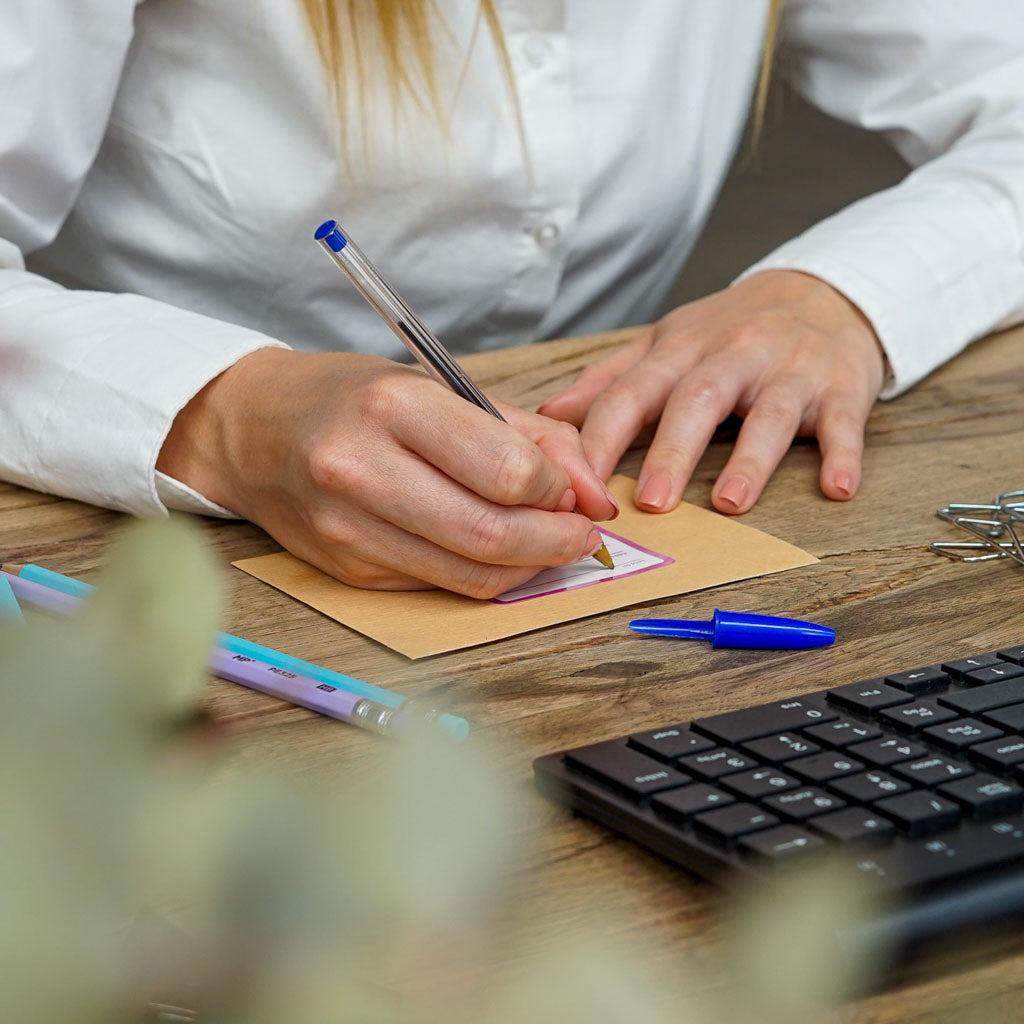 Close-up of a person's hands writing on a brown envelope with a blue pen, next to a black keyboard and colorful pens on a wooden desk. Ideal for showcasing personalized stationery or office supplies