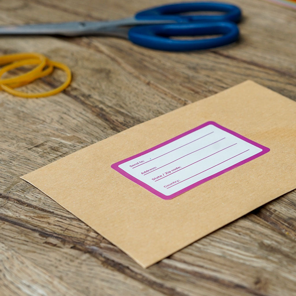 A brown envelope with a purple-bordered mailing label featuring fields for 'Send to:', 'Address:', 'State / Zip code:', and 'Country'. In the background, blue-handled scissors and yellow rubber bands are blurred on a wooden table, emphasizing office and shipping supplies.