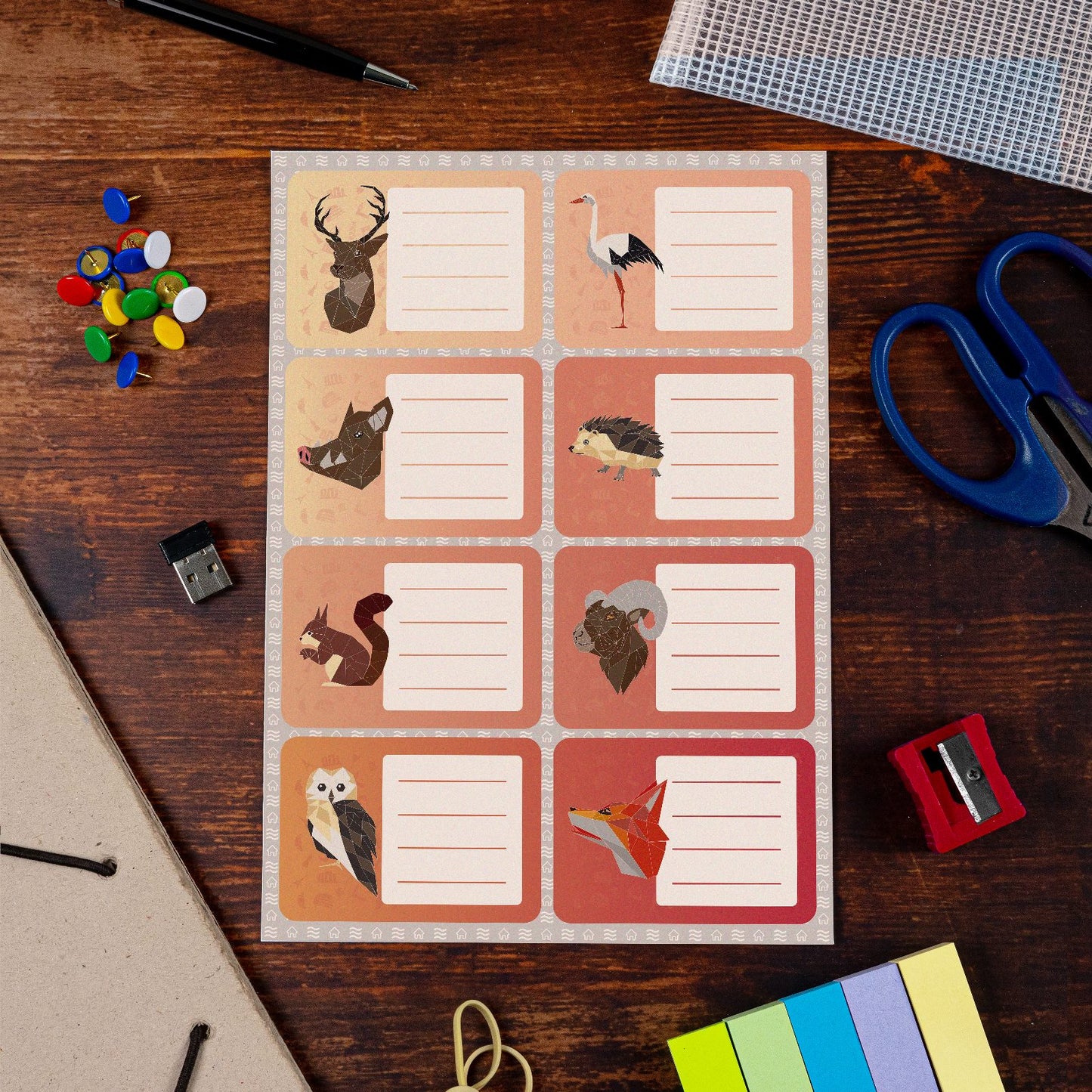 Sheet of rectangular animal-themed name labels on a wooden desk, surrounded by office supplies like scissors, pushpins, and sticky notes. The labels feature illustrations of a deer, stork, wild boar, hedgehog, squirrel, ram, owl, and fox, each with blank lines for writing. Ideal for personalizing belongings for school or home