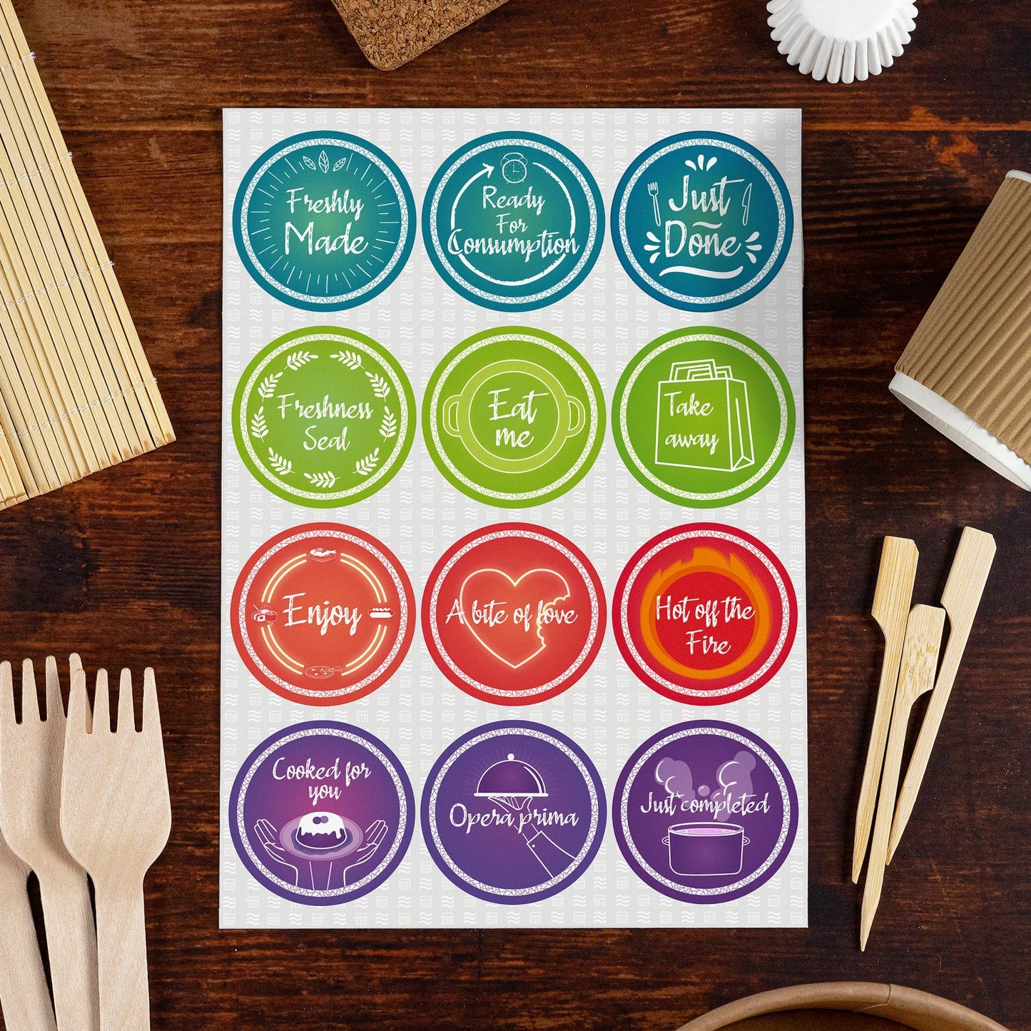 Sheet of colorful circular food preparation and serving labels on a wooden table, surrounded by bamboo utensils and food packaging accessories. Labels include 'Freshly Made,' 'Ready for Consumption,' 'Just Done,' 'Freshness Seal,' 'Eat me,' 'Take away,' 'Enjoy,' 'A bite of love,' 'Hot off the Fire,' 'Cooked for you,' 'Opera prima,' and 'Just completed,' perfect for restaurants, bakeries, or home meal prep.