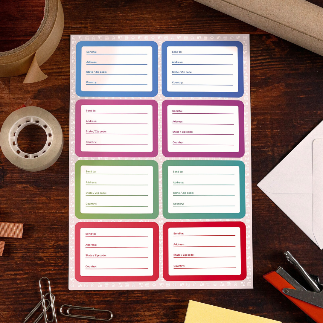 Sheet of colorful premium mailing labels on a wooden desk, surrounded by packing tape, clear tape, and office supplies. Features eight address labels in blue, pink, green, and red borders, ready for shipping and organization.