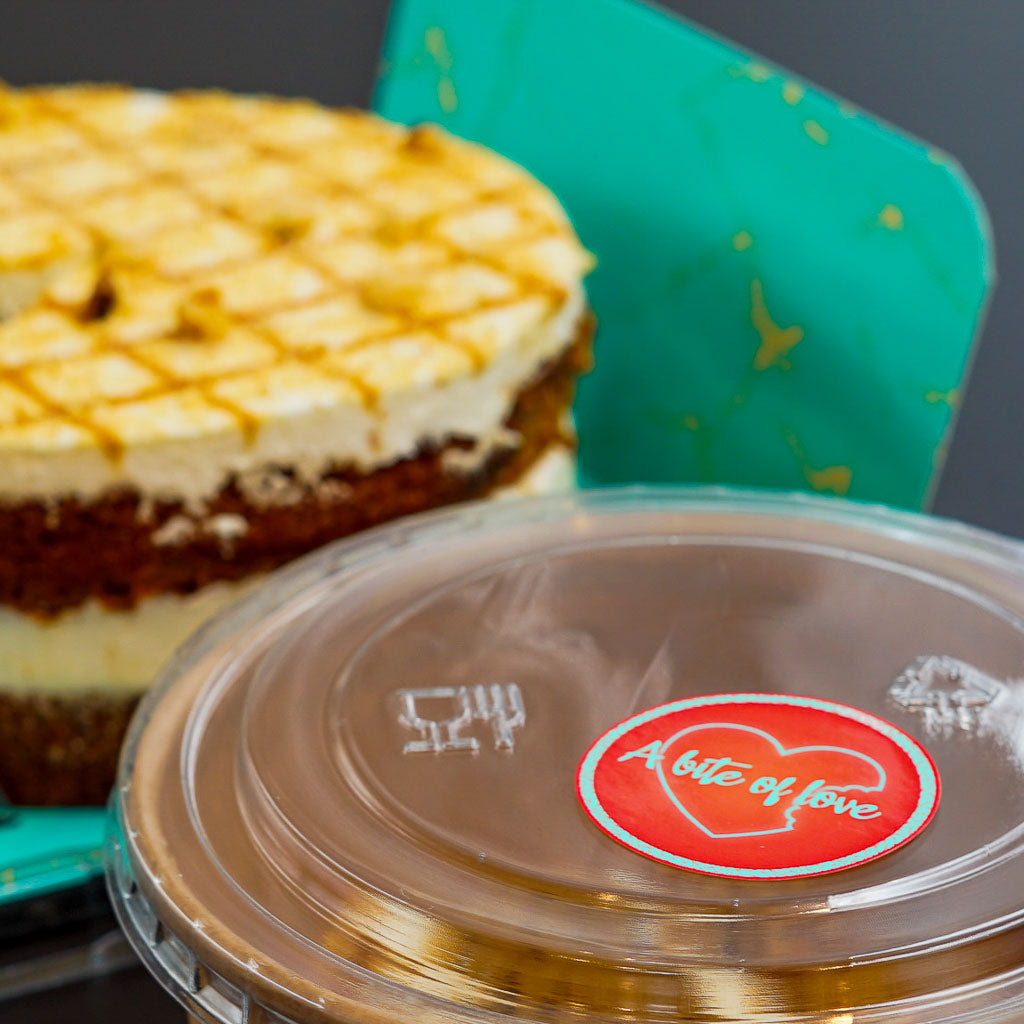 A clear plastic dessert container with a red oval 'A bite of love' sticker featuring a heart icon. In the blurred background, a delicious-looking carrot cake with white frosting and caramel drizzle is visible, suggesting a bakery or cafe offering sweet treats