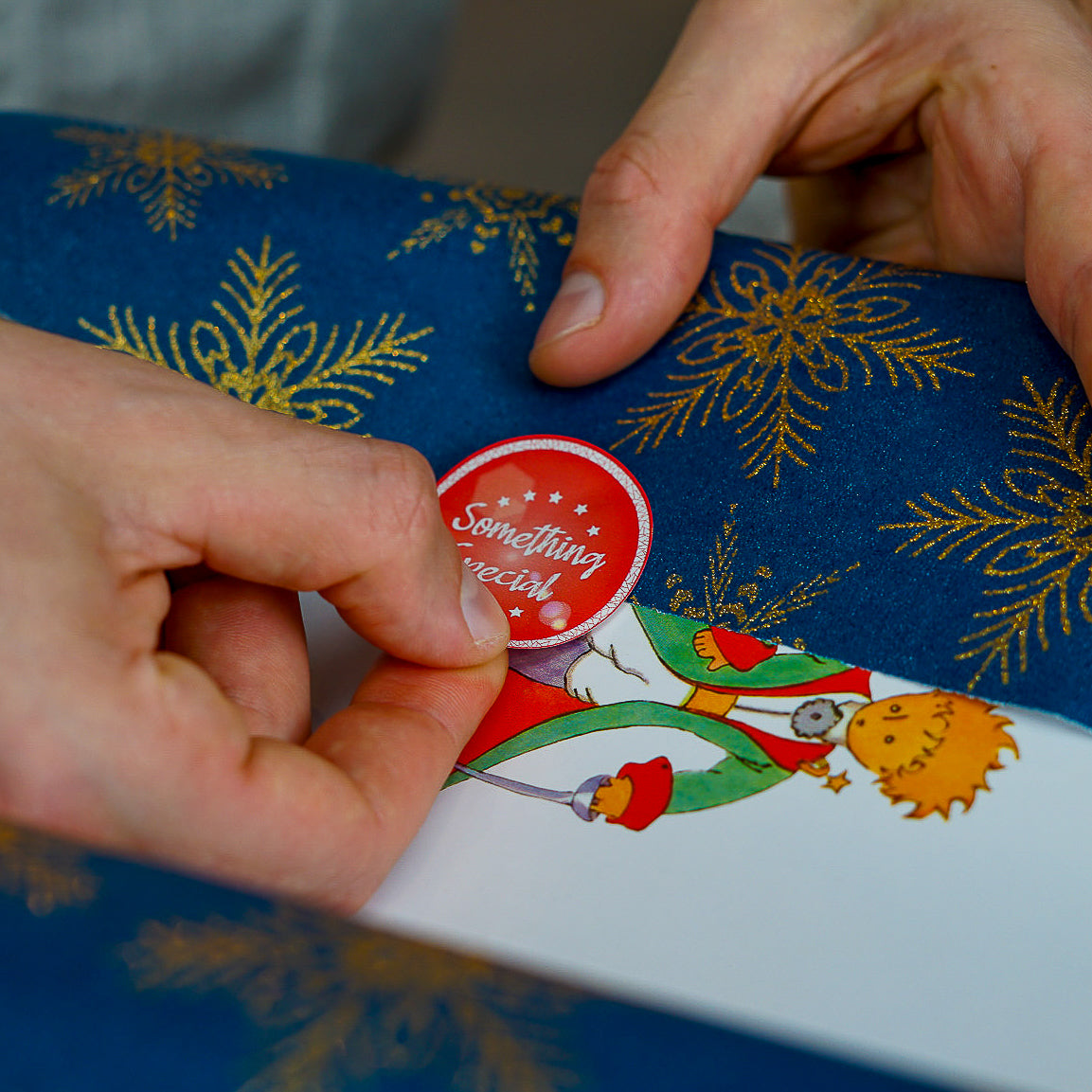 Hands placing a red circular 'Something special' sticker with stars onto a gift wrapped in blue paper with gold snowflakes and an illustration of The Little Prince. The sticker adds a personal touch to a thoughtful present.