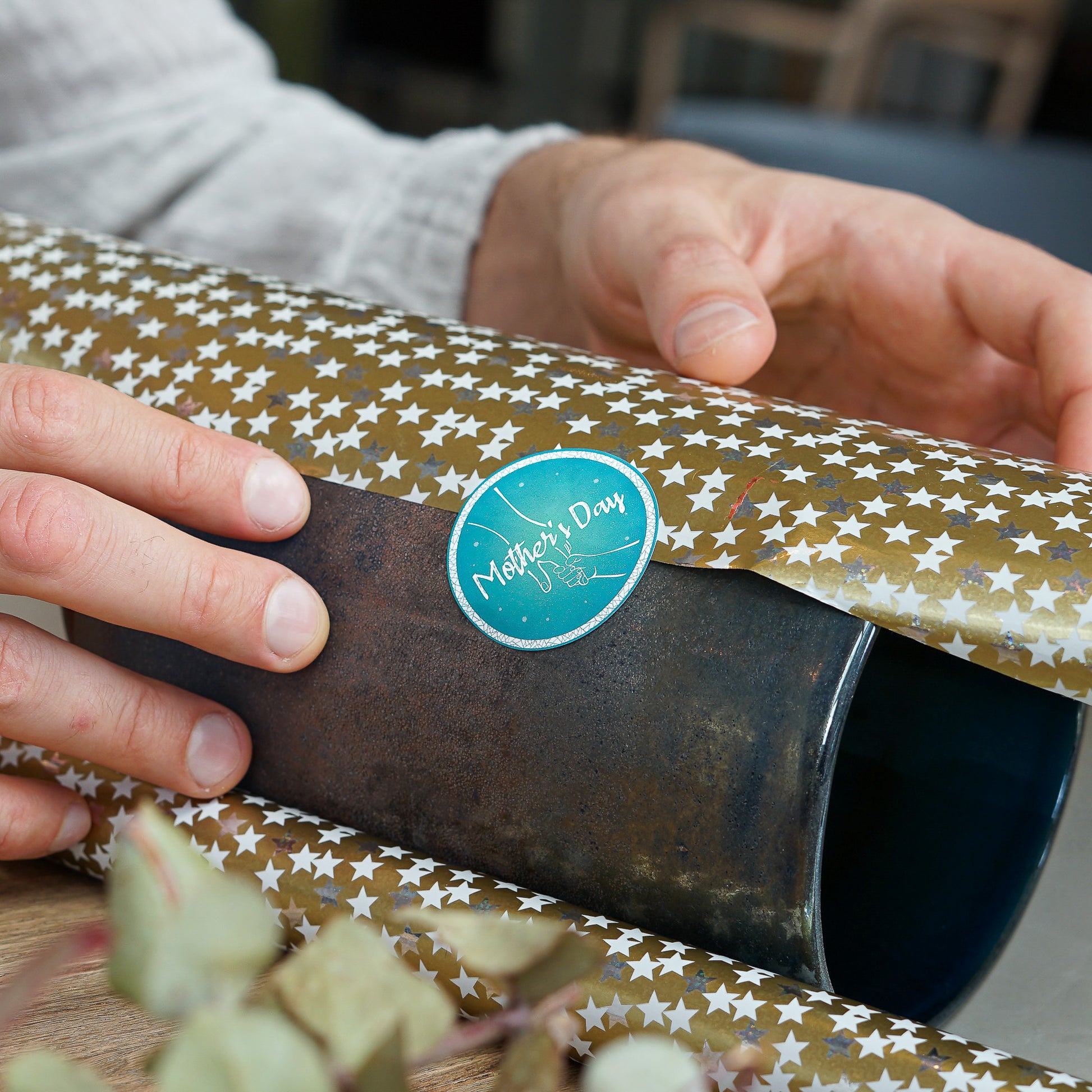 Hands wrapping a gift in gold star-patterned paper, with a teal circular 'Mother's Day' sticker prominently displayed on the package. The sticker features an outline of a mother and child's hands, perfect for adding a special touch to holiday gifts