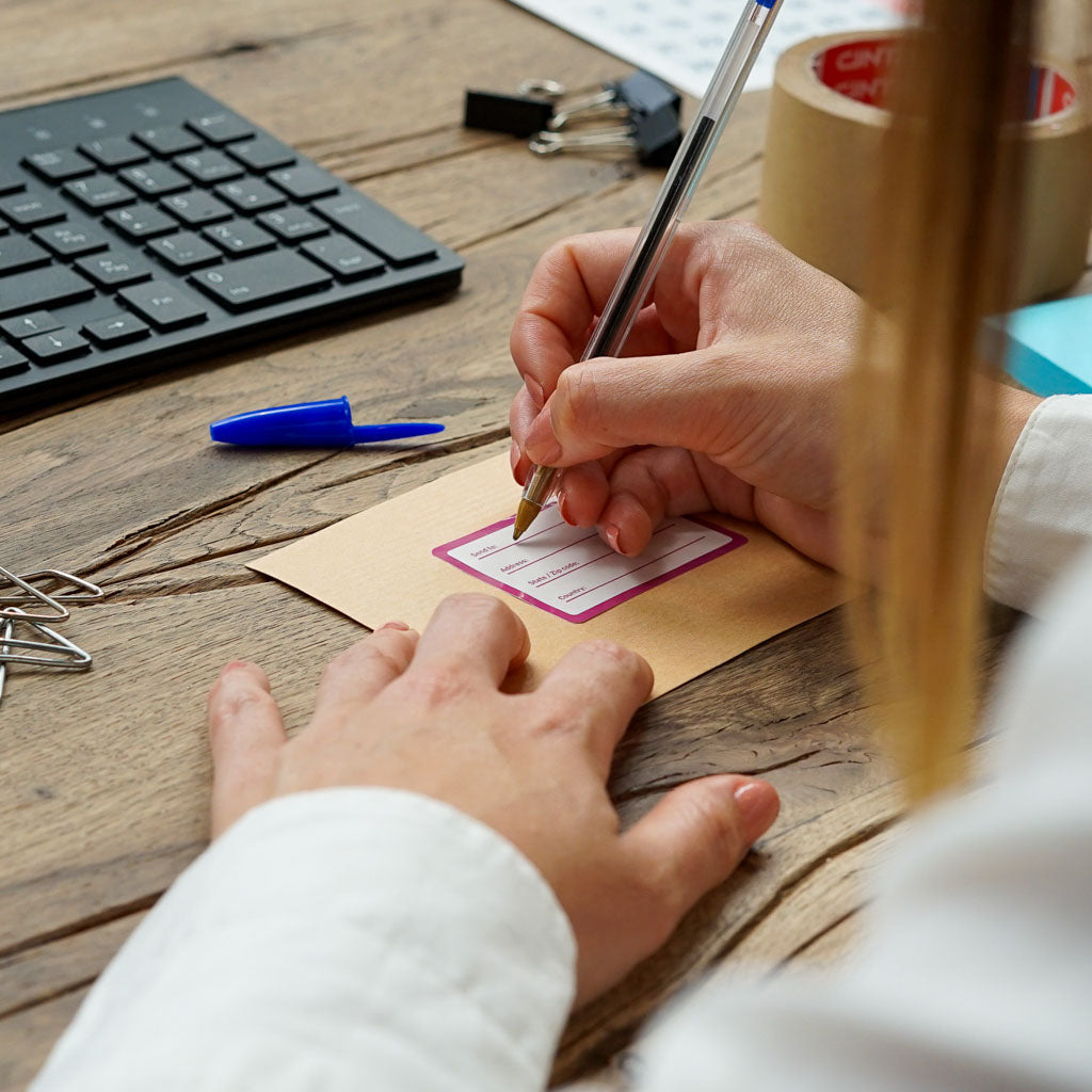 Overhead view of a person writing on a brown envelope with a purple-bordered mailing label, using a clear blue pen. A black keyboard, packing tape, and binder clips are visible on the rustic wooden desk, perfect for showcasing personalized shipping supplies.