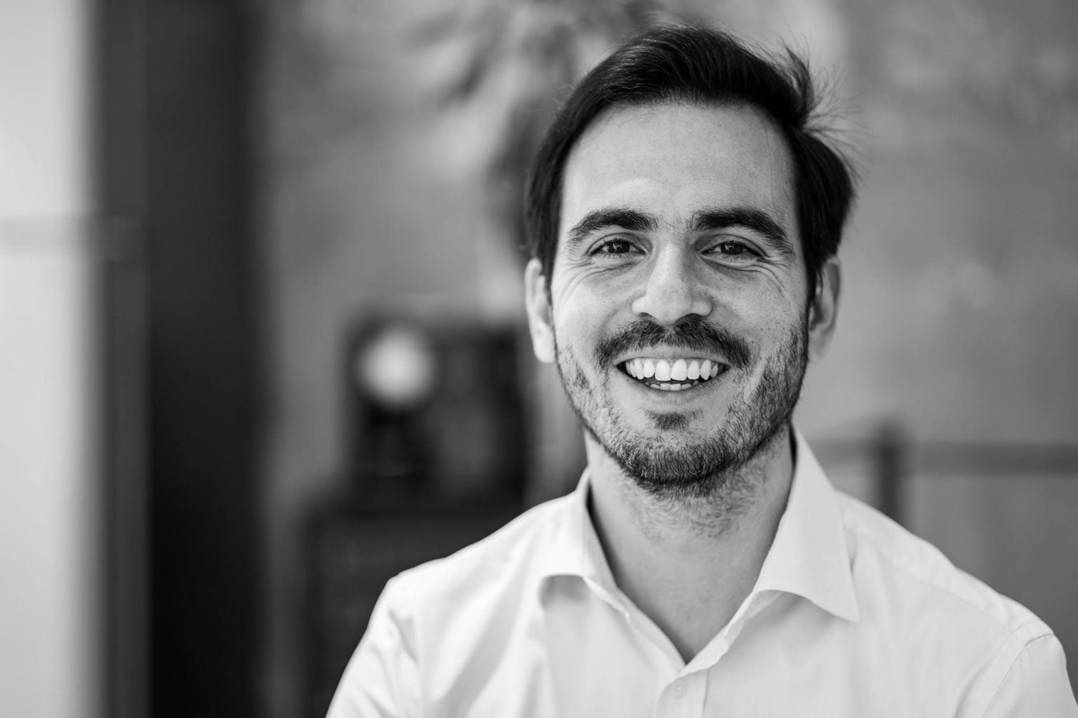 Black and white portrait of a smiling man with dark hair and a beard, wearing a light-colored collared shirt, looking directly at the camera. Professional headshot for a business profile or team page