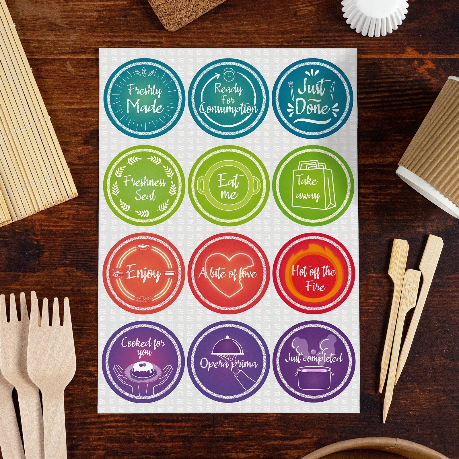 Sheet of colorful circular food preparation and serving labels on a wooden table, surrounded by bamboo utensils and food packaging accessories. Labels include 'Freshly Made,' 'Ready for Consumption,' 'Just Done,' 'Freshness Seal,' 'Eat me,' 'Take away,' 'Enjoy,' 'A bite of love,' 'Hot off the Fire,' 'Cooked for you,' 'Opera prima,' and 'Just completed,' perfect for restaurants, bakeries, or home meal prep.